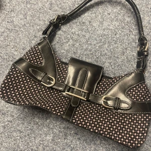 Svart och rosa axelväska med prickigt mönster vintage - Snygg axelväska med ett svart och rosa prickigt mönster. Väskan har svarta skinnliknande detaljer och två dekorativa spännen framtill. Den har en dragkedja upptill för säker förvaring. Perfekt för att ge din outfit en unik touch!