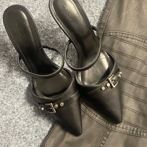 Kitten heels - Snygga svarta klackskor med spetsig tå och dekorativa spännen. Skorna har en elegant design med smala remmar och en stilren look. Perfekta för att ge en extra touch till din outfit.