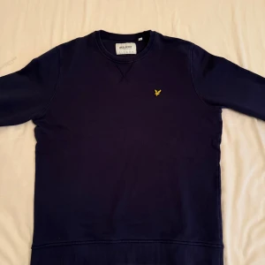 Lyle & Scott tröja - Säljer en stilren mörkblå tröja från Lyle & Scott med deras ikoniska logga på bröstet. Tröjan har långa ärmar och en rund halsringning. Perfekt för en avslappnad look. Använts en del därav fläcken på ryggen, syns inte så mycket i verkligheten är mest på bild med lampa