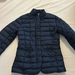 Mörkblå dunjacka från Gant - Snygg mörkblå dunjacka från Gant med quiltad design och knappar framtill. Jackan har en stilren krage och ett mönstrat innerfoder i blått och beige. Perfekt för kyligare dagar!