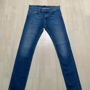 Blå jeans från Boss - Snygga blå jeans från Boss med klassisk femficksdesign och en läderpatch med logotyp på baksidan. De har en rak passform och är perfekta för en stilren look. Dragkedja och knapp framtill. Hugo Boss Delaware strl 33/34