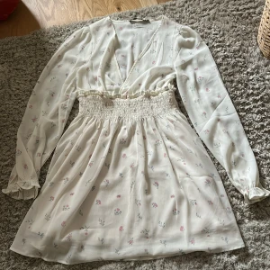 Blommig klänning från 157 - Söt blommig klänning från 157 i en ljus färg med små rosa och blå blommor. Klänningen har långa ärmar med volangdetaljer vid ärmsluten och en smockad midja för en fin passform. Perfekt för vår och sommar!