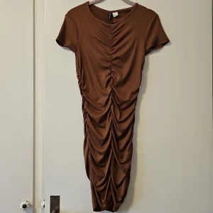 Brun klänning från H&M Divided - Snygg brun klänning från H&M Divided i en åtsittande modell med rynkade detaljer framtill och baktill. Klänningen har korta ärmar och är tillverkad i ett stretchigt material av polyester och elastan. Perfekt för en stilren look!