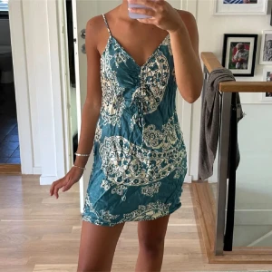 Mönstrad kortklänning med smala axelband - Snygg kortklänning i blått med ett unikt mönster och smala axelband. Perfekt för varma dagar och somriga kvällar. Klänningen har en v-ringad design och en åtsittande passform som framhäver figuren.