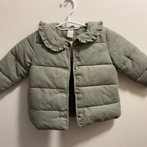 Grön quiltad jacka med volangkrage - Söt grön quiltad jacka från Zara med volangdetaljer runt kragen. Jackan har knappar framtill och är perfekt för de kallare månaderna. Den är v för små barn och ger både stil och värme.
