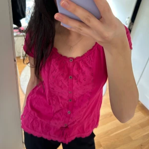 Rosa blus med knappar - Säljer en söt rosa blus med korta ärmar och knappar framtill. Blusen har ett fint broderat mönster och en liten volangdetalj vid fållen. Perfekt för en somrig look, tex till midsommar. 