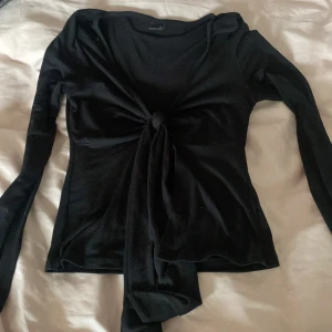 Svart långärmad knyt topp från Gina Tricot - Snygg svart långärmad knyt topp från Gina Tricot i storlek XS. Toppen har en knytning framtill som ger en cool look. Perfekt för att styla med jeans eller kjol.