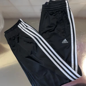 Svarta träningsbyxor från Adidas - Snygga svarta träningsbyxor från Adidas med de klassiska tre vita ränderna längs sidorna. Byxorna har en elastisk midja och är perfekta för sport och träning. Adidas-loggan är tryckt på framsidan.