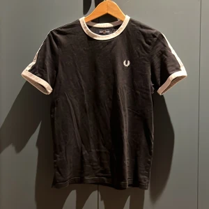Svart t-shirt från Fred Perry - Snygg svart t-shirt från Fred Perry med vita detaljer på ärmar och krage. Den har en klassisk passform och det ikoniska Fred Perry-logotypen broderad på bröstet. Perfekt för en stilren look.