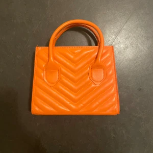 Orange  handväska - Snygg orange handväska med quiltat mönster och praktiska handtag. Väskan har en dragkedja för säker förvaring och en avtagbar axelrem för extra flexibilitet. Perfekt för att liva upp din outfit med en färgklick!