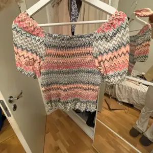 Säljer en färgglad topp från KappAhl med zigzag-mönster i rosa, blått och vitt. Toppen har korta puffärmar och en elastisk passform som ger en smickrande siluett. Perfekt för en somrig look! 🌸