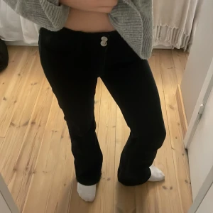 Svarta byxor från Gina Tricot - Snygga svarta byxor från Gina Tricot med bootcut-stil. Byxorna har två knappar framtill och bakfickor med en diskret broderad detalj. Perfekta för en stilren look.