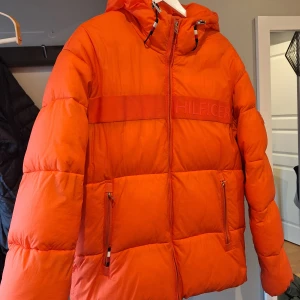 Orange pufferjacka från Tommy Hilfiger - Säljer en snygg orange pufferjacka från Tommy Hilfiger. Jackan har en dragkedja framtill och två praktiska fickor med dragkedjor. Perfekt för att hålla värmen under kyliga dagar. Den har en huva för extra skydd mot väder och vind. Den har en liten fläck, syns knappt. Är skonsamt nytvättad . Skickar bild vid intressen. 