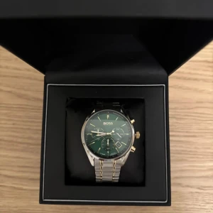 Grön klocka från Boss - Elegant klocka från Boss med grön urtavla och guldfärgade detaljer. Armbandet är i silverfärgat metall och klockan levereras i en stilren svart box. Perfekt för den som vill ha en tidlös accessoar.