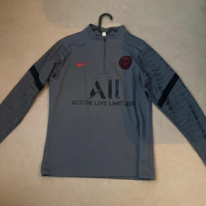 Psg tröja - Säljer en grå träningsjacka från Nike med PSG-logga och texten 'Accor Live Limitless' på framsidan. Jackan har en halv dragkedja och långa ärmar med svarta detaljer. Perfekt för fotbollsträning eller som en sportig vardagslook. Självklart är det inga skador priset kan alltid diskuteras först till kvarn