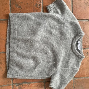 Grå stickad tröja från Zara - Säljer en stilren grå tröja från Zara. Den är kortärmad och har en mjuk, stickad känsla med ribbade kanter vid ärmslut och nederkant. Andvänt några fåtals gånger! 