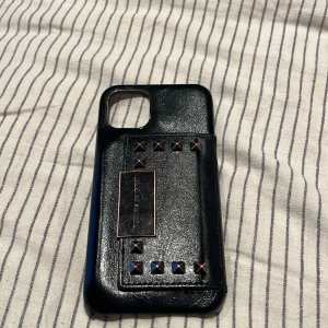 Svart mobilskal med kortfack från Ideal of Sweden - Snyggt svart mobilskal från Ideal of Sweden med praktiskt kortfack. Skalet har en stilren design med nitar och en insida i beige mocka. Perfekt för att hålla dina kort säkra och lättillgängliga.