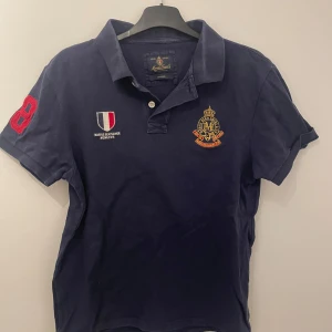 Mörkblå pikétröja från Morris Gent's - Säljer en snygg mörkblå pikétröja från Morris Gent's med broderade detaljer. Tröjan har en klassisk krage och korta ärmar. Perfekt för en stilren look. Skriv om ni har frågor och priset är inte hugget i sten!