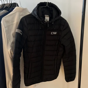 Svart dunjacka från ICANIWILL - Säljer en svart dunjacka från ICANIWILL i storlek XS. Jackan har en huva och dragkedja framtill. Perfekt för kyliga dagar med sin quiltade design och långa ärmar. Märkesdetaljer på ärmen och bröstet ger en sportig look.
