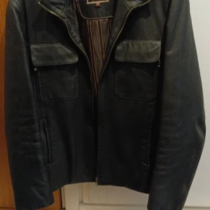 Svart skinnjacka från RZJ International - Snygg svart skinnjacka från RZJ International med dragkedja och två bröstfickor med lock. Jackan har en klassisk stil och är perfekt för en cool look. Den är långärmad och har en stilren design som passar till många olika outfits. Använt 2 gånger