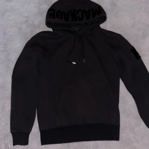 Svart hoodie från Mackage - Säljer en stilren svart hoodie från Mackage med tryck på huvan och ärmen. Den har en praktisk känguruficka och justerbar dragsko i huvan. Perfekt för en avslappnad look.