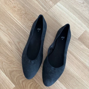 Oanvända svarta ballerinaskor från H&M - Säljer ett par stilrena svarta ballerinaskor från H&M i storlek 36. Perfekta för en enkel och bekväm look. Skorna har en klassisk design med rundad tå och är tillverkade i syntetmaterial.