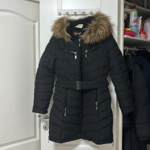 Svart dunjacka med pälsdetaljer - Snygg svart dunjacka med avtagbar päls på huvan och dragkedja framtill. Jackan har ett bälte i midjan för en figursydd passform och flera praktiska fickor med dragkedjor. Perfekt för kalla dagar.