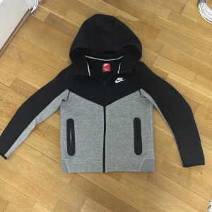 Grå och svart huvtröja från Nike - Säljer en grå och svart huvtröja från Nike med dragkedja och huva. Tröjan har långa ärmar och praktiska fickor med dragkedjor. Perfekt för en sportig look.