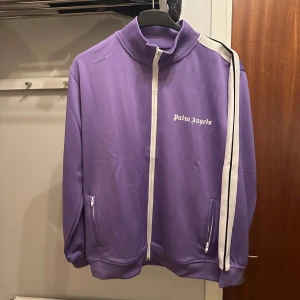 Lila träningsjacka från Palm Angels - Säljer en snygg lila träningsjacka från Palm Angels med vita detaljer och dragkedja. Jackan har långa ärmar med vita ränder längs sidorna och en hög krage. Säljer av hela garderoben då kläderna inte används längre.