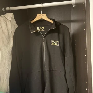 Svart hoodie från EA7 - Snygg svart hoodie från EA7 med dragkedja och logga på bröstet. Perfekt för en avslappnad stil. Den har en klassisk passform och är tillverkad i ett mjukt material. Har även en luva!