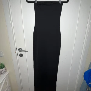 Svart axelbandslös klänning från Bershka - Snygg svart axelbandslös klänning från Bershka i en elegant lång modell. Perfekt för en stilren look med en smickrande passform. Klänningen har en slits på sidan för extra rörelsefrihet. Perfekt för att skapa en chic outfit!