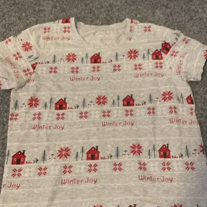 Grå t-shirt med vintermotiv - Mysig grå t-shirt med vintertema. Den har ett charmigt mönster med röda stugor, snöflingor och texten 'Winter Joy'. Perfekt för att sprida vinterkänsla.