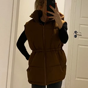 Brun pufferväst med dragkedja och stor huva - Säljer en snygg brun pufferväst med dragkedja och huva. Västen har en normal passform och är perfekt för kyligare dagar. Den är stilren och enkel att matcha med olika outfits. Lappen är borta men passar stlk M-L