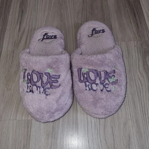 Lila tofflor med text - Mysiga lila tofflor med texten 'Love' broderad på ovansidan. Perfekta för att hålla fötterna varma hemma. Mjukt material och halkfri sula.