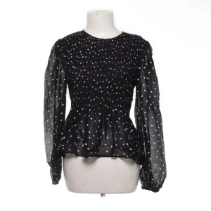Svart prickig blus från Zara - Säljer en elegant svart blus från Zara med vita prickar. Blusen har långa ärmar med volangdetaljer vid midjan. Storlek M