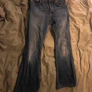 Blå jeans från Nudie Jeans - Säljer ett par klassiska blå jeans från Nudie Jeans med orange sömmar och snygg broderad detalj på bakfickorna. Perfekta för en avslappnad stil. De har en knappgylf och är tillverkade i slitstarkt denim. Bootcut. W30 L34 men känns mindre.