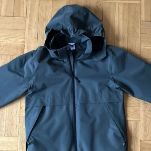 Mörkgrön regnjacka med huva McKinley - Snygg mörkgrön regnjacka med huva och dragkedja framtill. Jackan har långa ärmar och praktiska fickor med dragkedja. Perfekt för regniga dagar och håller dig torr och bekväm. 10 000 mm i vattenpelare.