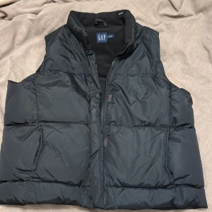 Svart dunväst från GAP - Säljer en svart dunväst från GAP i storlek M. Västen har en dragkedja framtill och är perfekt för kyligare dagar. Den är stilren och enkel att matcha med olika outfits.