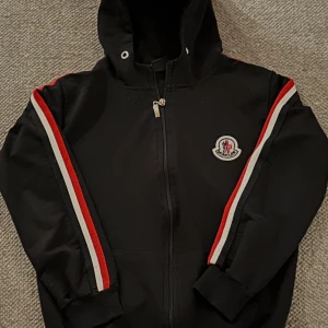 Svart huvtröja från Moncler - Säljer en svart huvtröja från Moncler med dragkedja och röd-vita ränder längs ärmarna. Tröjan har en broderad logotyp på bröstet och ribbade muddar vid ärmslut. Perfekt för en stilren look.