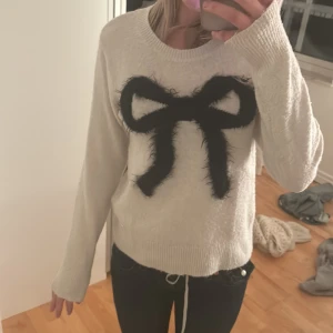 Vit tröja med svart rosett - Säljer en mysig beige tröja med en stor svart rosett på framsidan. Tröjan har långa ärmar och en rund halsringning. Perfekt för en stilren och söt look.