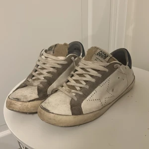 Golden Goose sneakers - Säljer ett par Golden Goose sneakers i vitt och grått med stjärndetalj på sidan. Du får med kvitto, box och dustbag. Skorna har snörning och en vintage-look med slitna detaljer. Perfekta för en avslappnad stil. Priset är inte hugget i sten!!!
