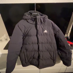 Svart dunjacka från Adidas - Snygg svart dunjacka från Adidas med huva och dragkedja. Ny pris är 1200 men den passar inte längre och den har legat i mitt rum ett tag utan användning. Det är inga problem med jackan och den är väldigt varm
