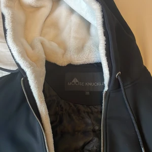 Svart jacka med huva från Moose Knuckles - Snygg svart jacka från Moose Knuckles (äkta, kvitto finns) med mjukt foder och huva. Jackan har dragkedja framtill och ribbade muddar vid ärmslut och nederkant. Perfekt för kyligare dagar.  Slitna muddar tyvärr men toppenskick resten av jackan.