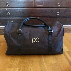 Brun weekendväska från Dolce & Gabbana - Snygg brun weekendväska från Dolce & Gabbana med svarta handtag och logotypdetaljer. Perfekt för kortare resor eller som en stilren gymväska. Väskan har en dragkedja för säker förvaring.
