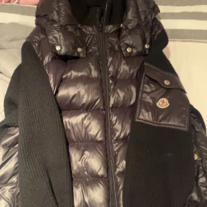Svart pufferjacka från Moncler - Snygg svart pufferjacka från Moncler med dragkedja och knappar. Jackan har en quiltad design och en hög krage för extra värme. Perfekt för kyliga dagar. Dragkedjan gick sönder så vi har bytt den till en vanlig 