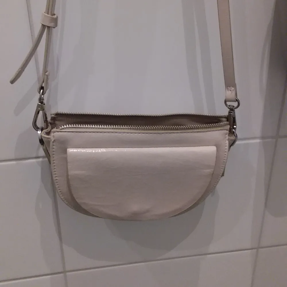 Snygg beige axelväska i syntetmaterial med justerbar axelrem. Väskan har en praktisk dragkedja upptill och en stilren design som passar till många tillfällen.. Laukut & Käsilaukut.