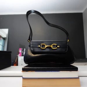 Svart axelväska med gulddetaljer - Snygg svart axelväska med gulddetaljer. Väskan har en elegant design med ett dekorativt guldfärgat spänne på framsidan. Perfekt för att ge en stilren touch till din outfit.