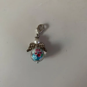 Ängelhänge med blommönster - Charmigt ängelhänge med silverfärgade vingar och en pärla som huvud. Kroppen är en glaskula med ett färgglatt blommönster i rosa och blått. Perfekt som ett unikt smycke eller dekoration.