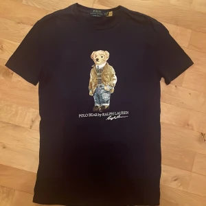Marinblå t-shirt med Polo Bear från Ralph Lauren XS - Snygg svart t-shirt från Ralph Lauren med det ikoniska Polo Bear-motivet på framsidan. T-shirten har en slim fit passform och är kortärmad, perfekt för en stilren look. Bra skick och knappt använd. Storlek XS. Ny pris 1000kr