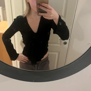 Svart v-ringad topp - Snygg svart v-ringad topp med knytning framtill. Toppen har långa ärmar och en figurnära passform, perfekt för en stilren look. Från designbysi 